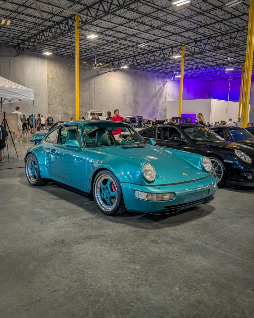 Teal Porsche 911 coupe in indoor car show with people and other vehicles in background
