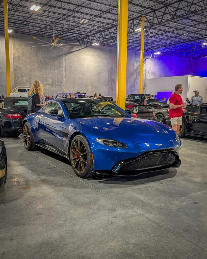 Blue Aston Martin sports car front 3/4 parked indoors at a car event with people and other vehicles in background