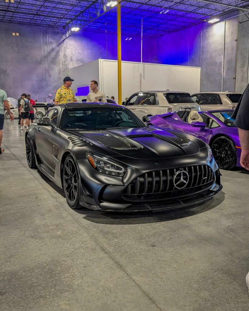 Black Mercedes-Benz AMG GT parked indoors with a purple convertible and silver SUVs in the background.