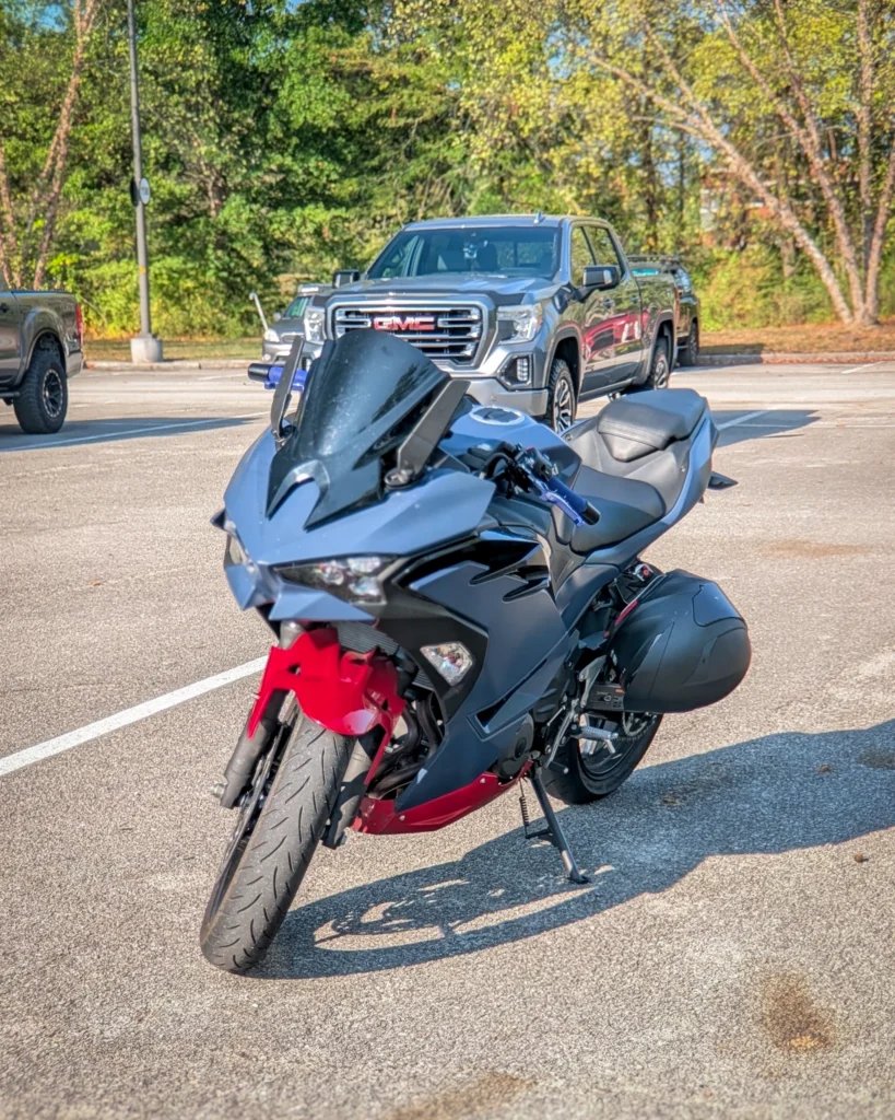 Black and red Kawasaki Ninja 500 in a parking lot with a gray GMC pickup truck in the background among trees