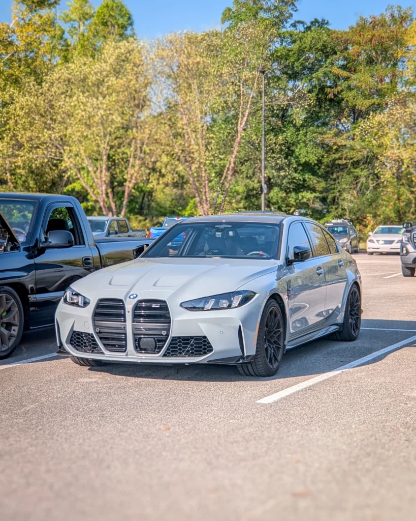 Silver BMW M3 sedan parked in a lot with other vehicles and trees in the background under clear sky