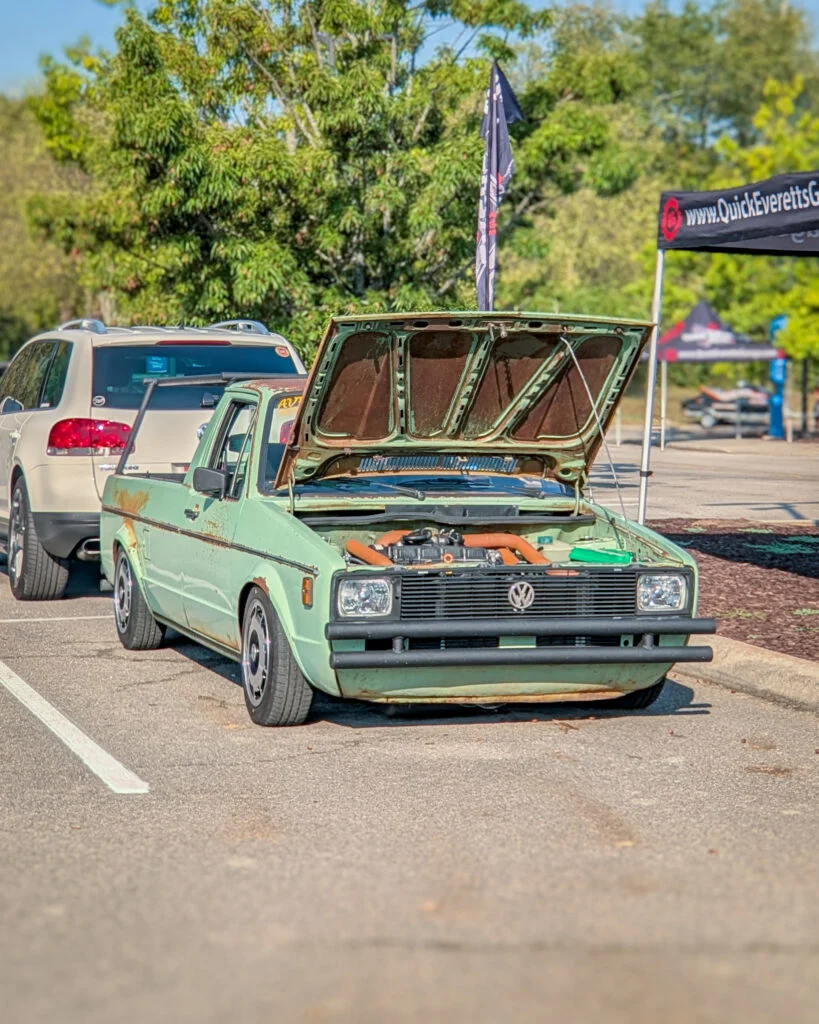 A light green Volkswagen Rabbit Pickup sits with its hood open in a parking lot at an outdoor car event.