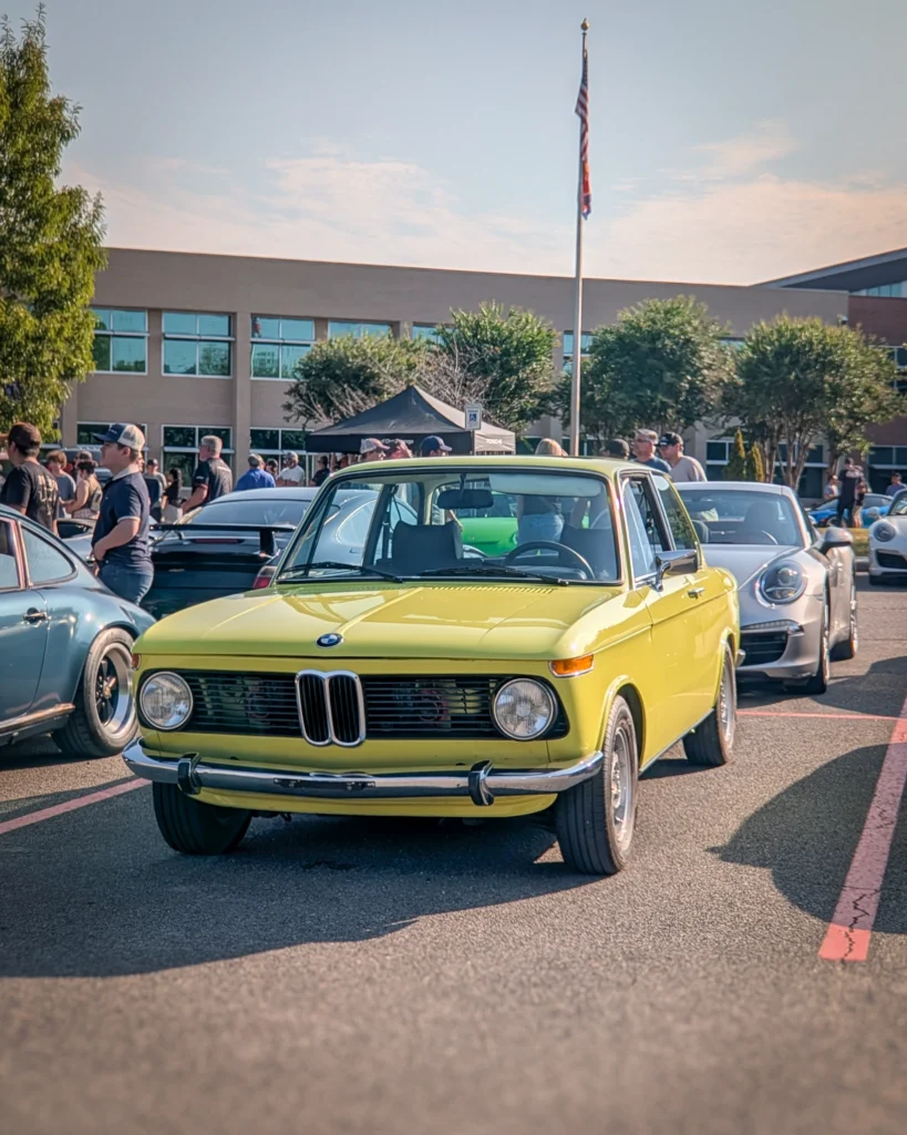 Yellow BMW 2002 parked in lot with a silver Porsche 911 and a crowd at an outdoor car event