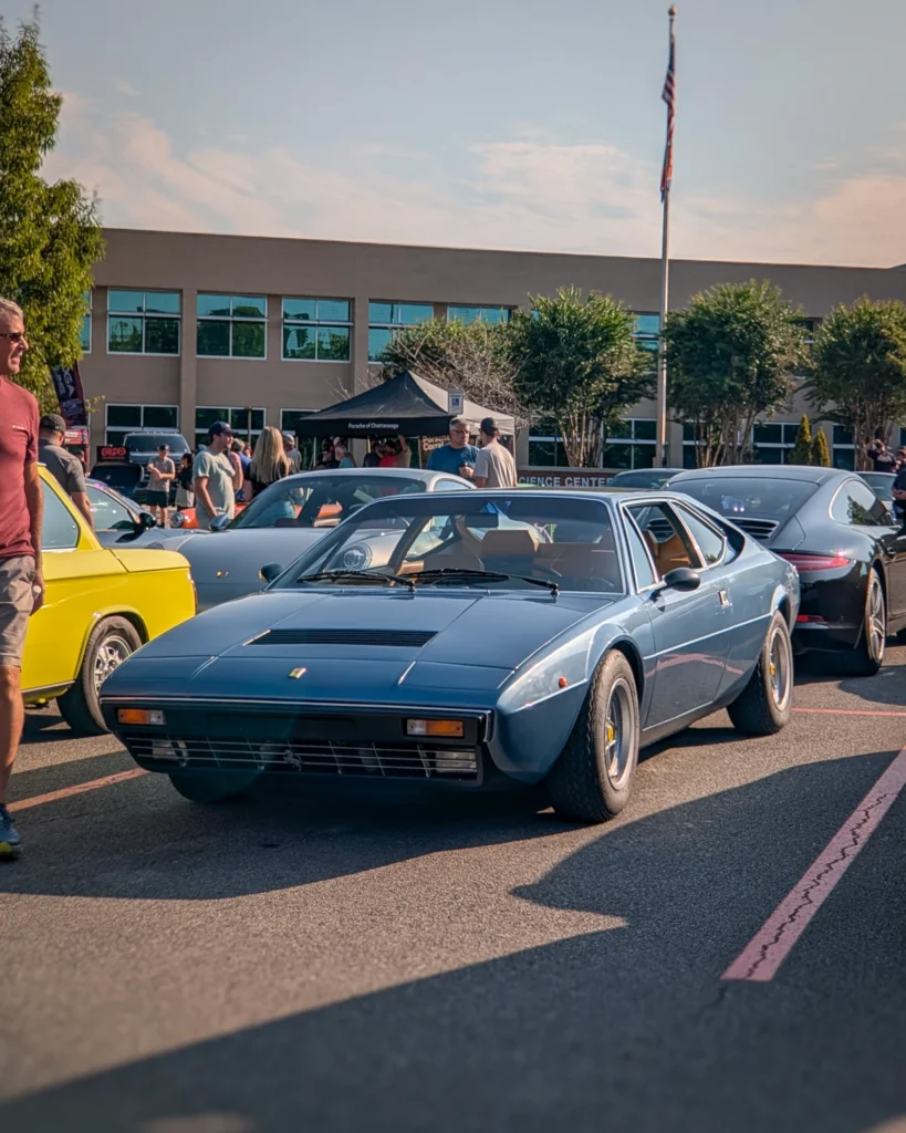 Blue Ferrari Dino 308 GT4 coupe in a parking lot at a car event with other vehicles and people in the background.