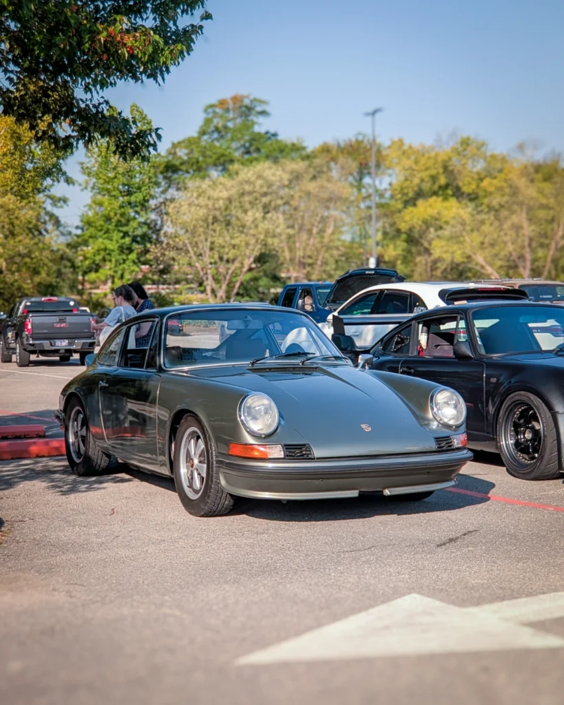 Green Porsche 911 coupe parked in a lot with other vehicles and trees in the background under clear sky.