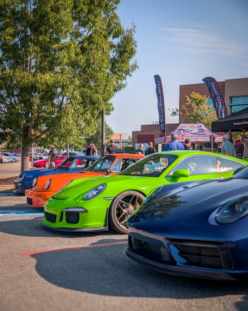 Row of Porsche sports cars in blue, green, orange, and blue parked outdoors with people and trees in background