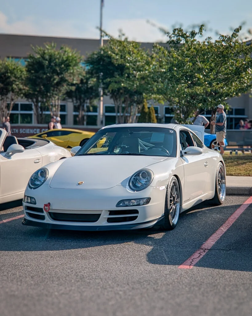 White Porsche 911 parked at a car event with other vehicles and people in the background