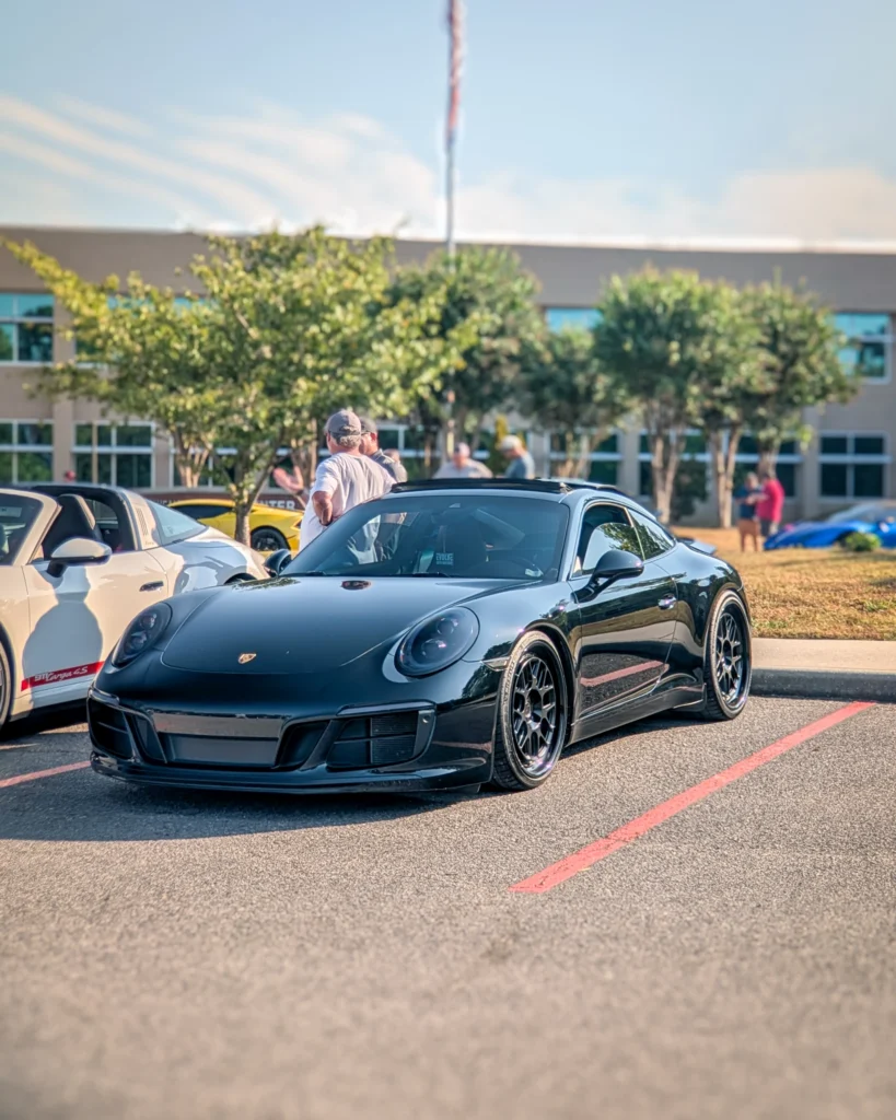 Black Porsche 911 coupe parked at a car event with other vehicles and people in the background