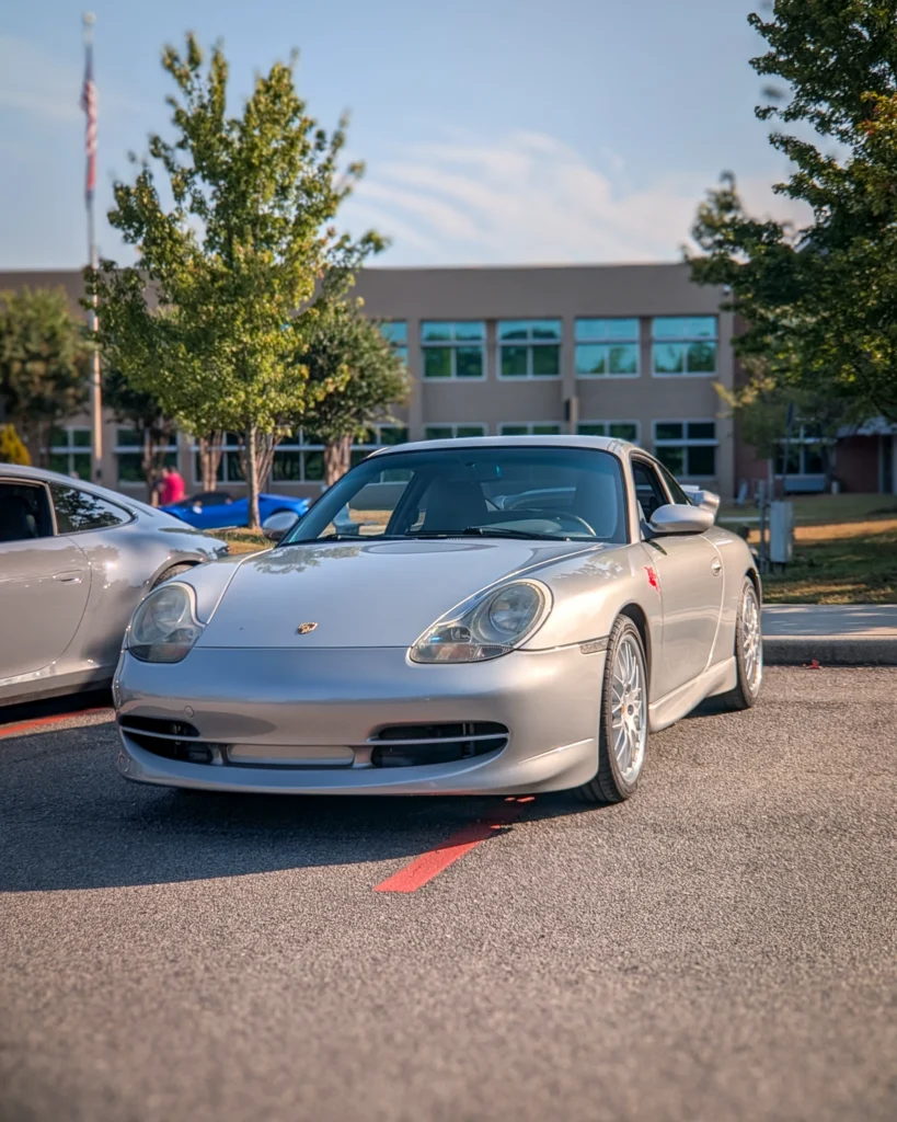 Silver Porsche 911 parked outdoors with building, trees, and another silver car nearby on a clear day.