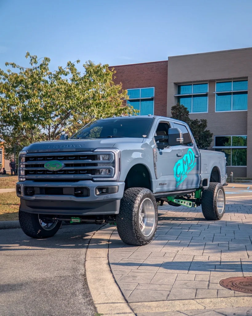 Silver Ford Super Duty pickup truck with custom lift and wheels parked by curb in front of brick building under blue sky.