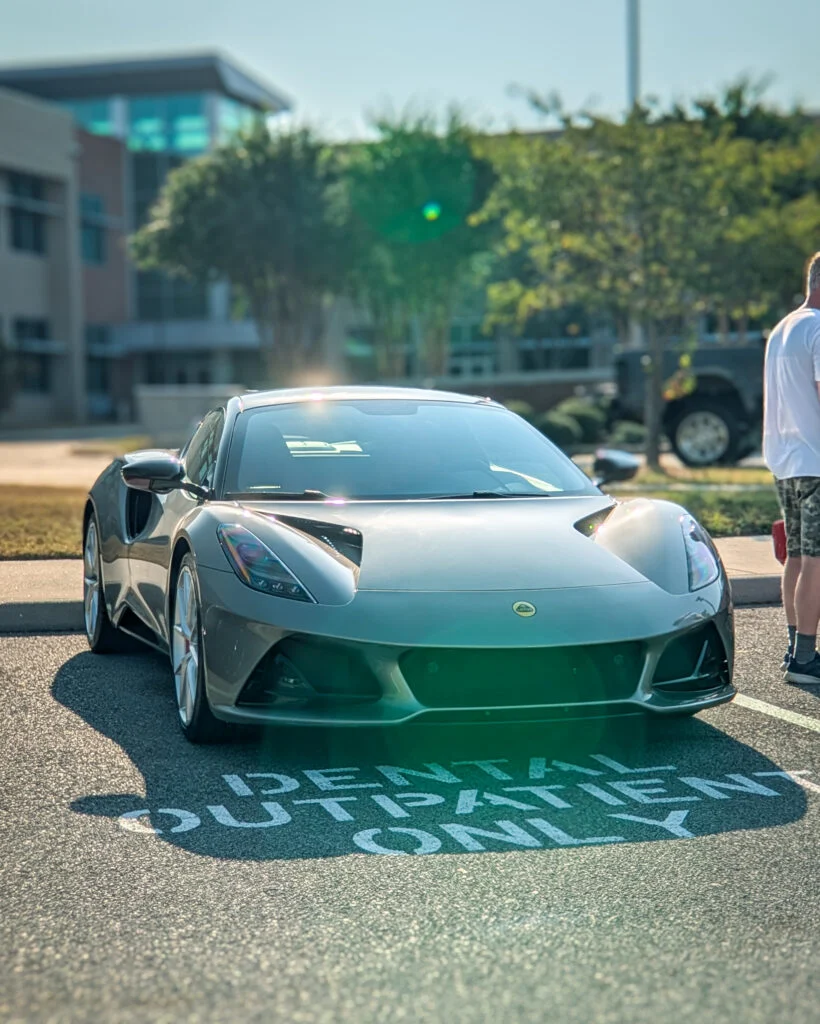 A silver Lotus Emira is parked outside a building, viewed from the front three-quarter angle.