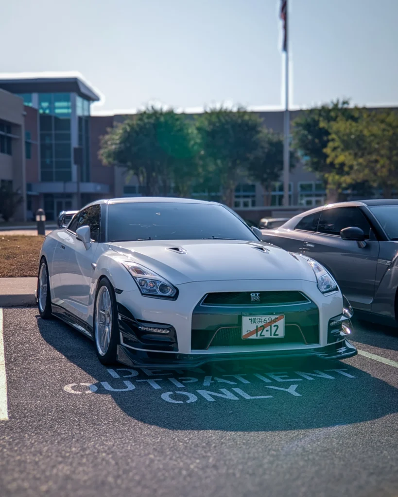 White Nissan GT-R parked in a designated space near a building and adjacent to a gray Nissan GT-R under clear sky