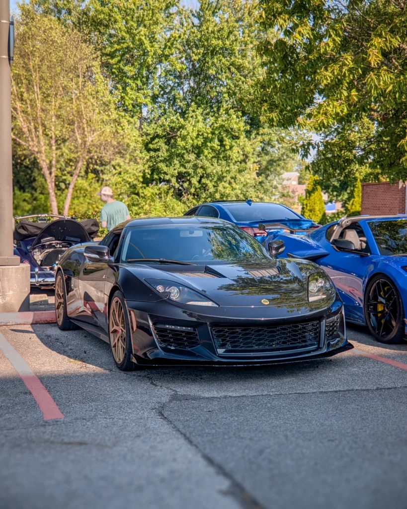 A black Lotus Evora is parked among other cars beneath trees at an outdoor car event.