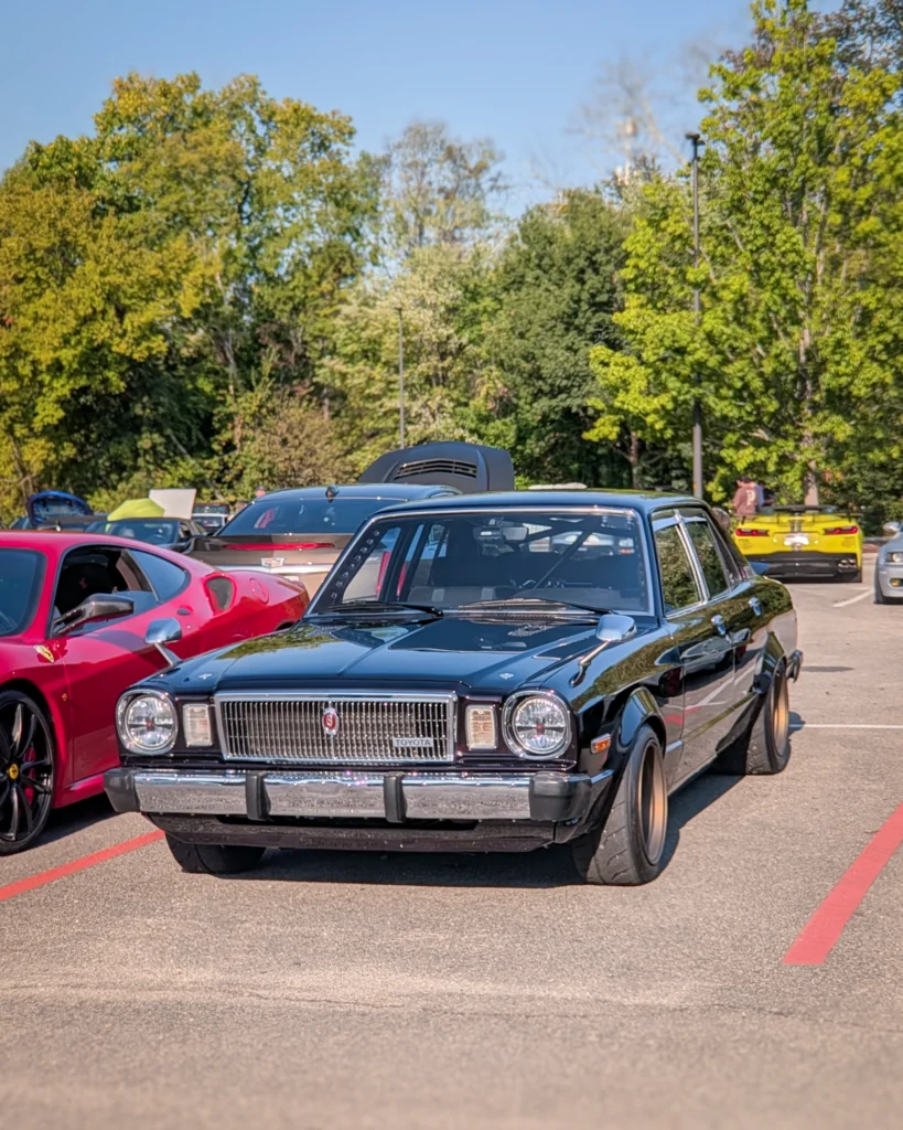 Black Toyota Cressida parked at a car event with a red Ferrari coupe and other cars in background under clear skies.