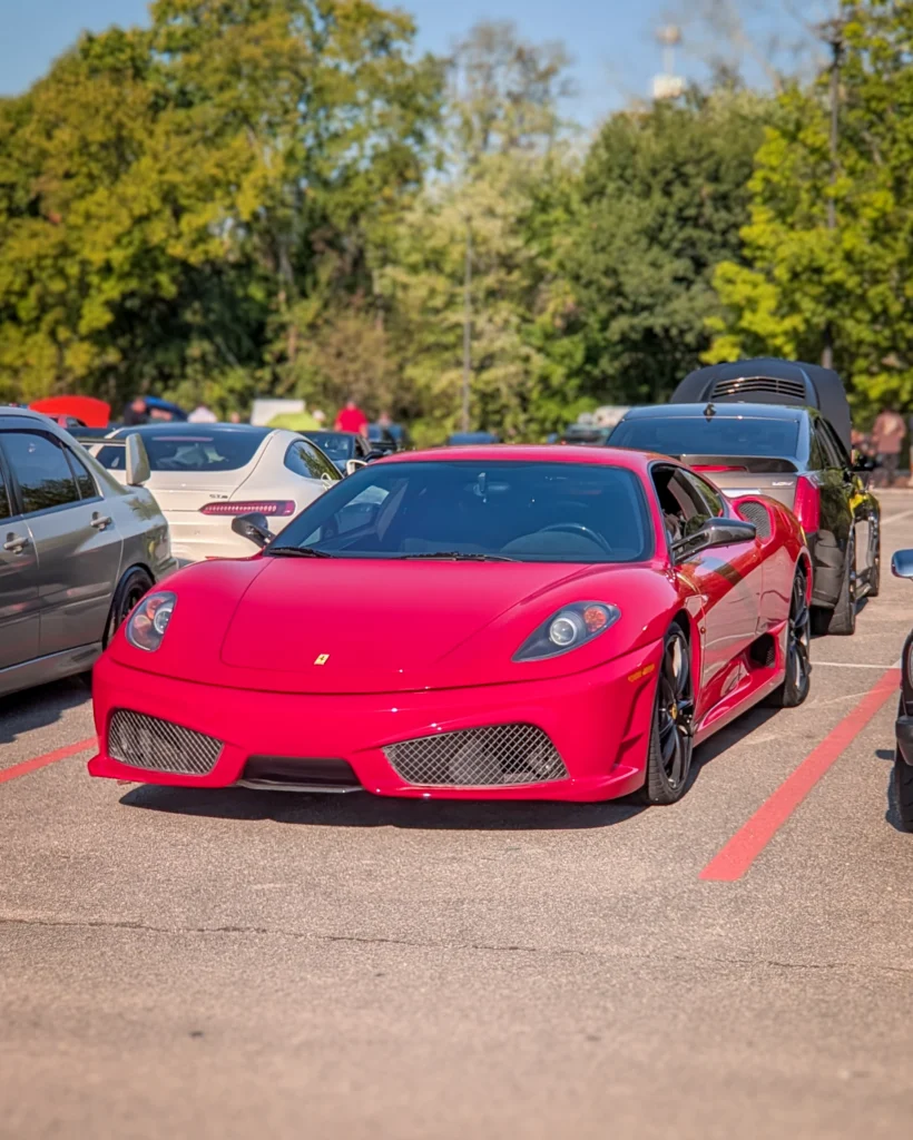 Red Ferrari F430 parked between other vehicles with a white Porsche 911 GT3 visible behind it at an outdoor car event.