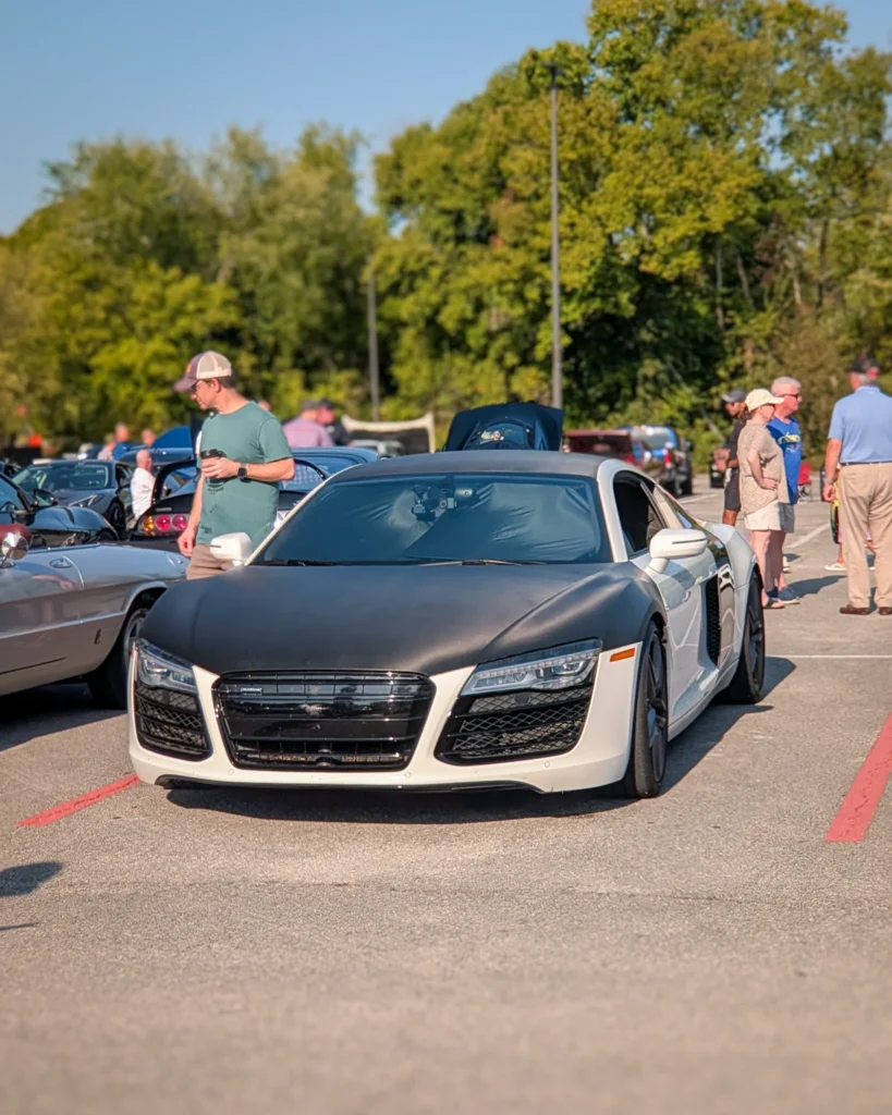 White Audi R8 with black hood and roof parked at an outdoor car event with people nearby and trees in background