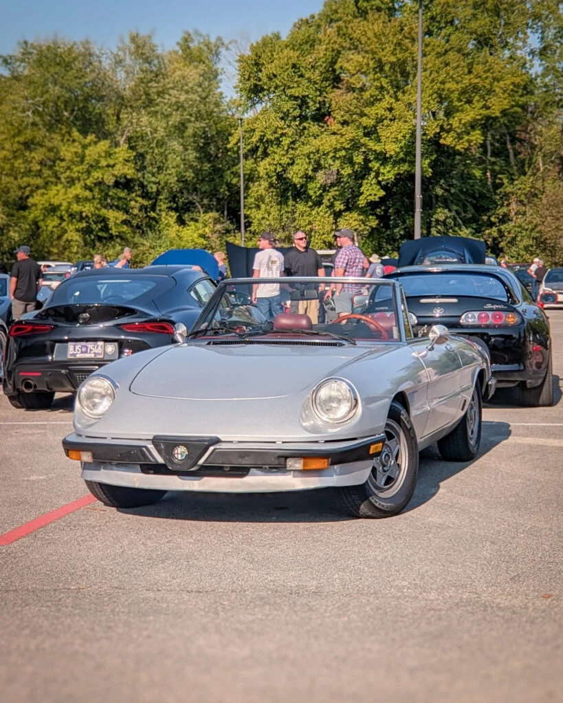 Silver Alfa Romeo Spider convertible parked among other cars at a daytime car meet with trees in the background