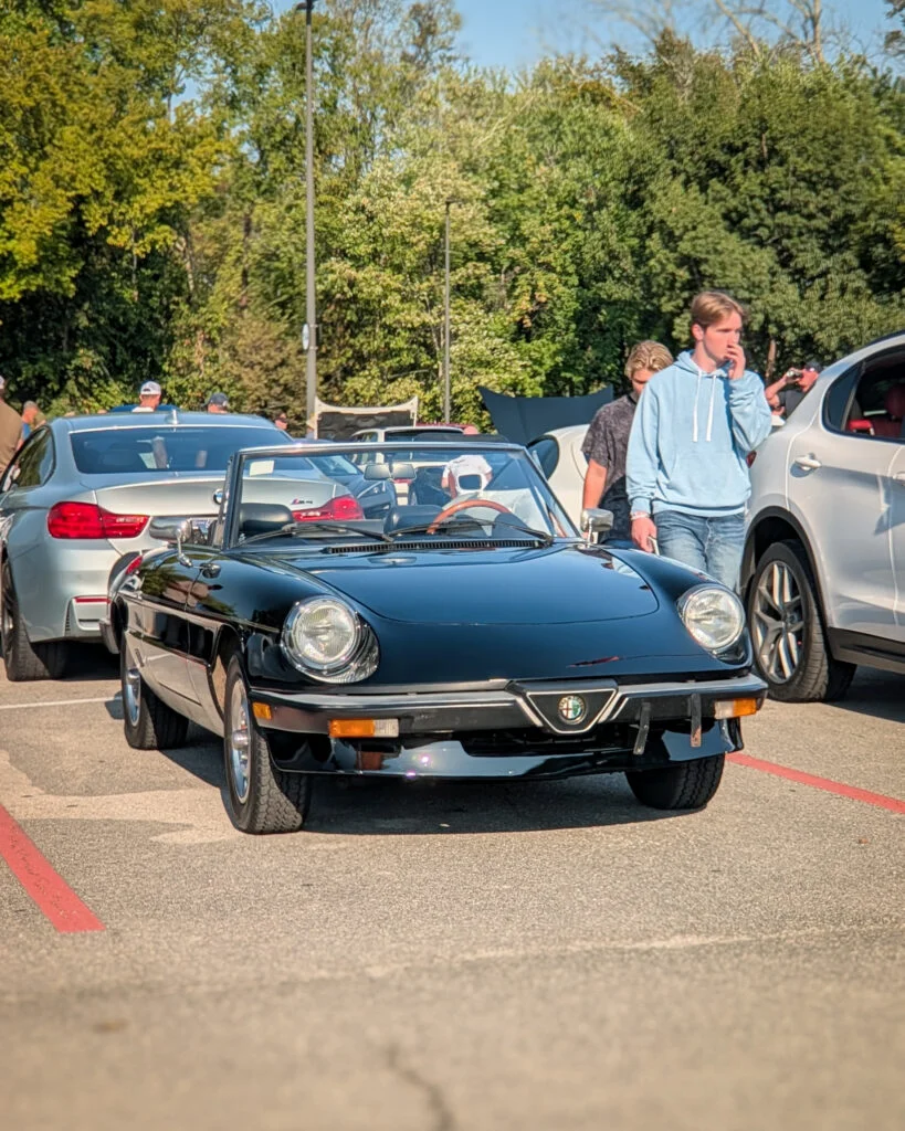 A black Alfa Romeo Spider with its top down is parked among cars and attendees at an outdoor car meet.