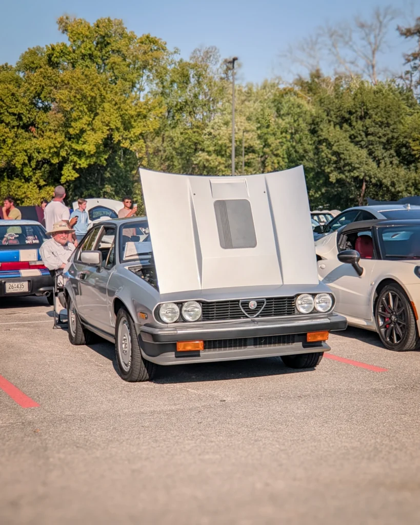 A silver Alfa Romeo GTV6 sits with its hood raised at an outdoor car show, with attendees and other cars nearby.