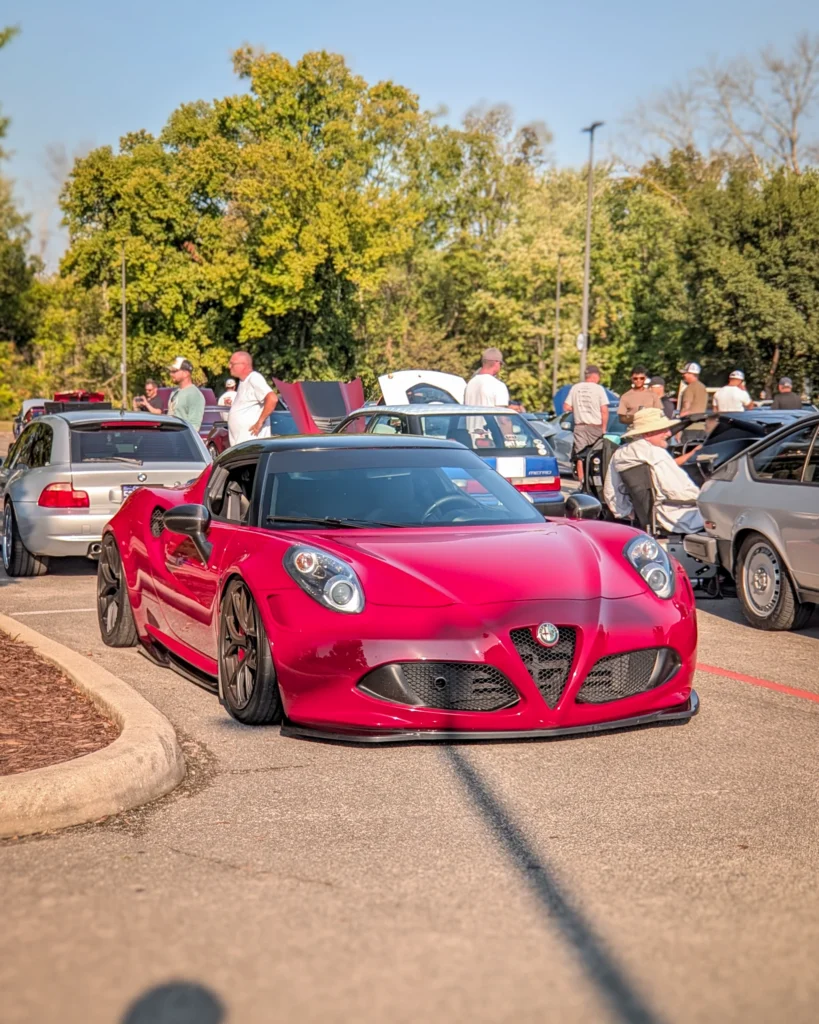 A red Alfa Romeo 4C parked at an outdoor car show with attendees and other cars behind it.