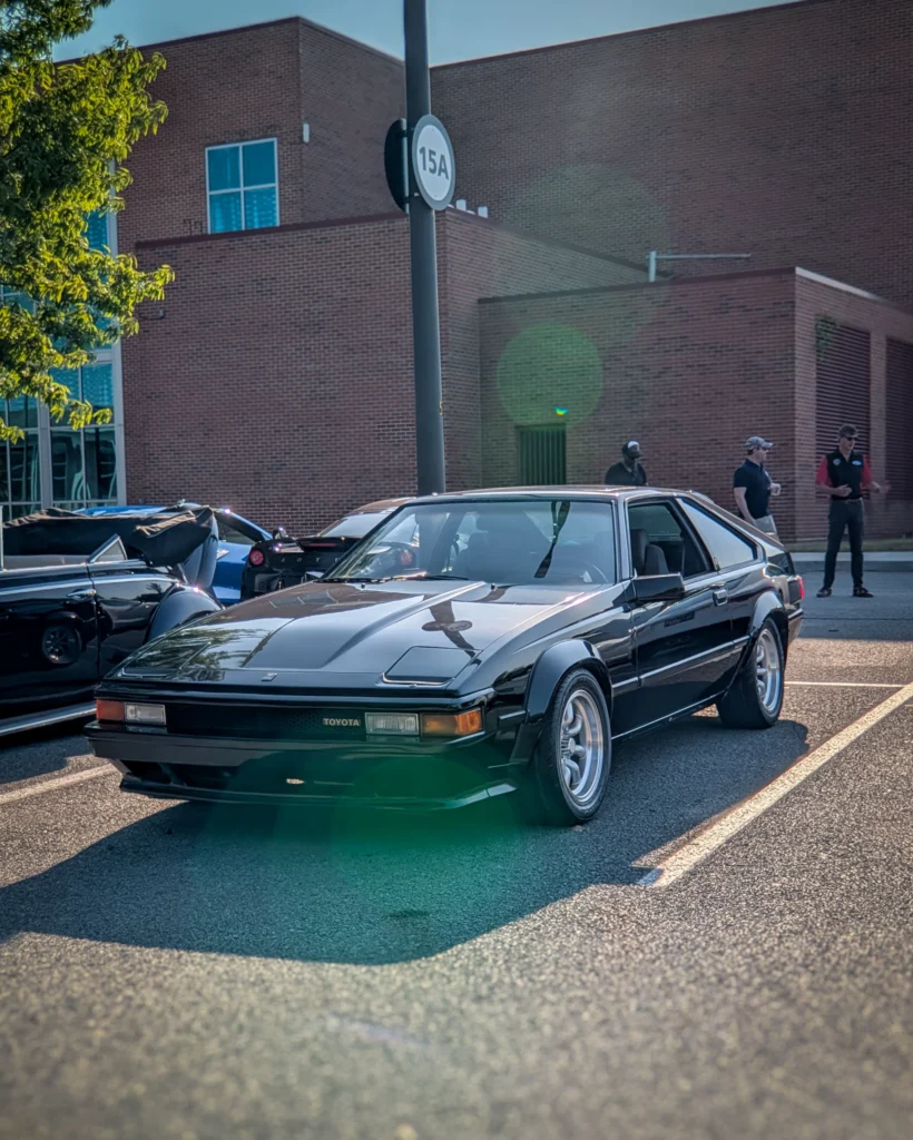 Black Toyota Supra coupe parked in a lot near a brick building with people standing nearby.