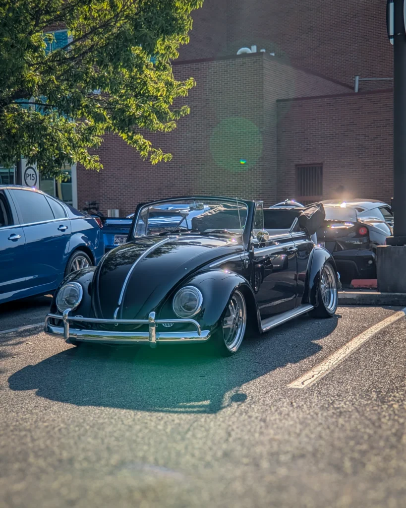 Black Volkswagen Beetle convertible parked in a lot near a brick building with other cars visible