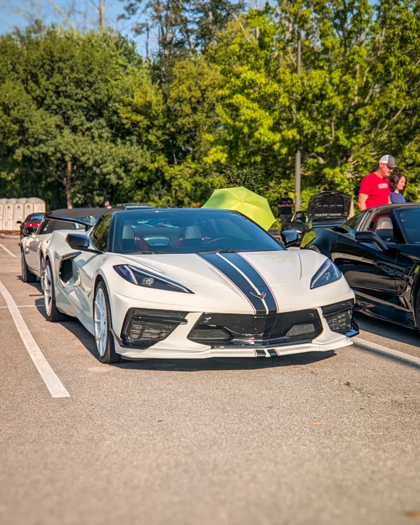 White Chevrolet Corvette with black racing stripes parked at a car event with other vehicles and people, surrounded by green