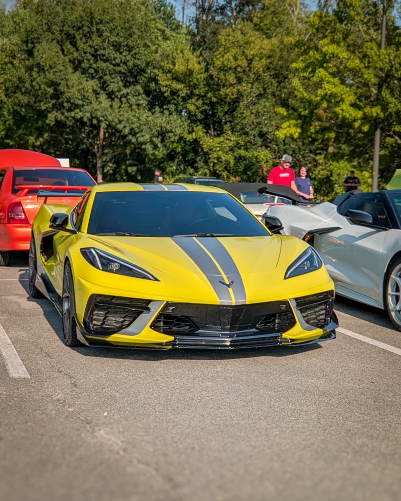 Yellow Chevrolet Corvette with black racing stripes parked in a lot among other cars and people in background