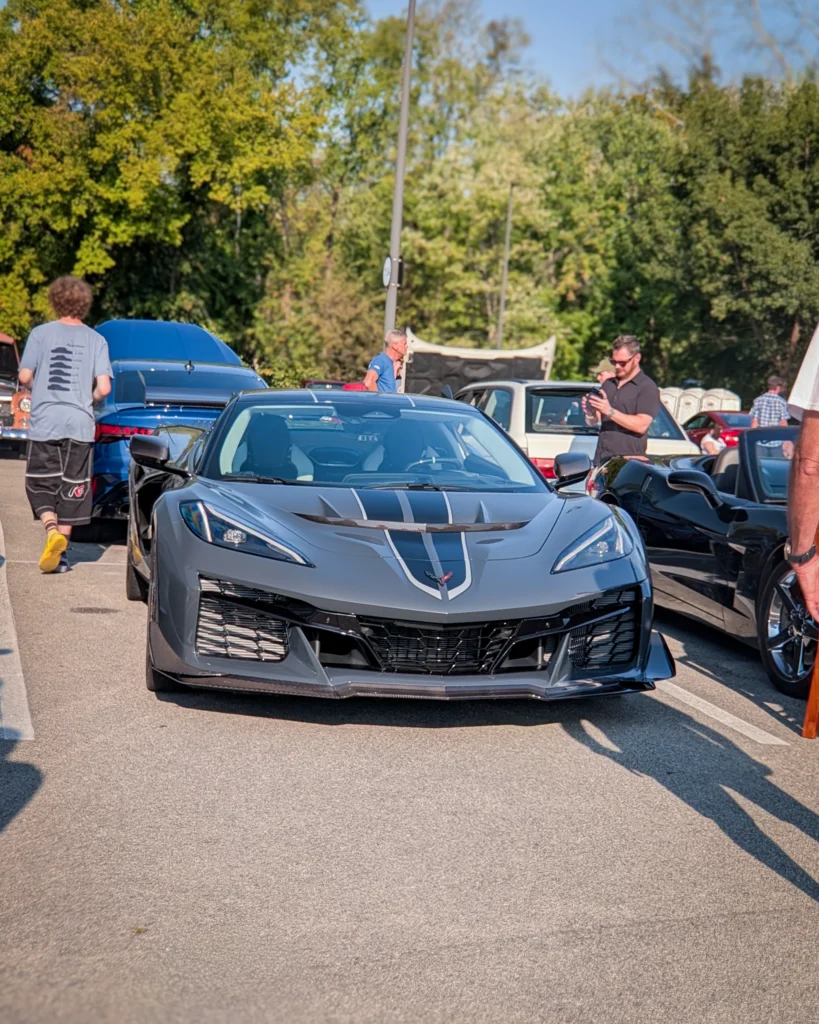 Gray Chevrolet Corvette with black racing stripes parked at an outdoor car event with people around and other cars visible
