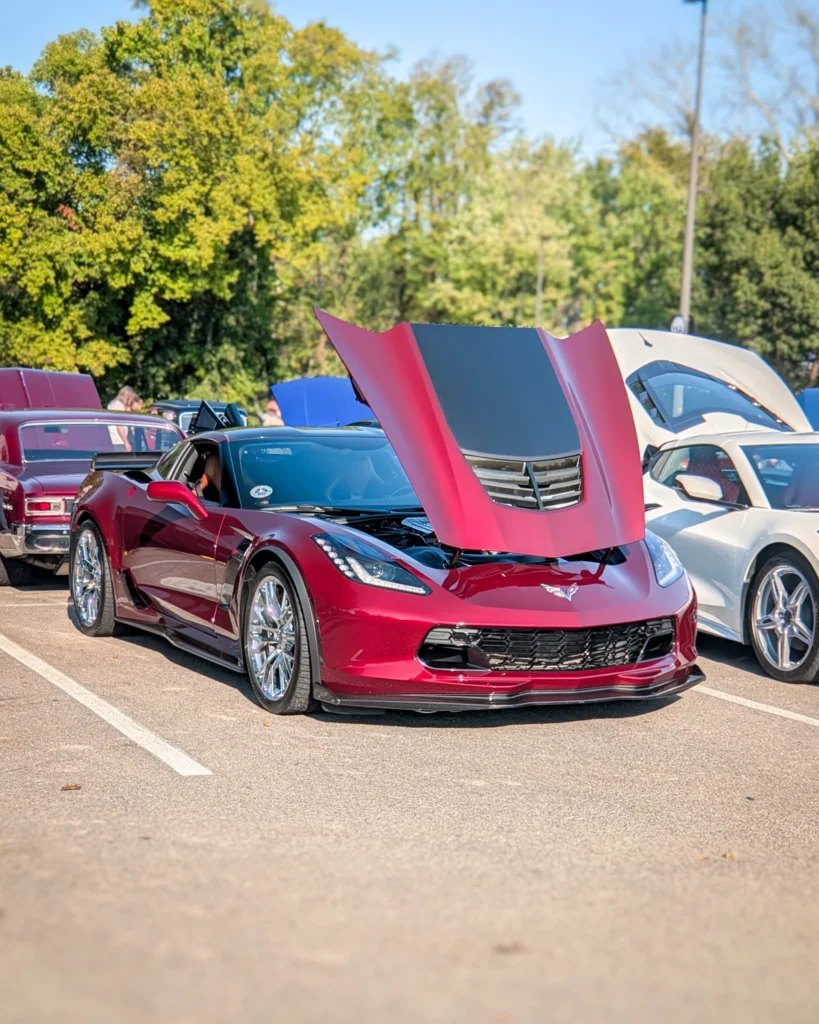 Red Chevrolet Corvette with raised hood parked next to a white Chevrolet Corvette with raised hood at an outdoor car event.