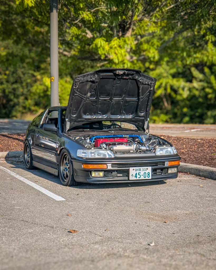 Dark-colored Honda CRX with its hood open parked in a lot near trees