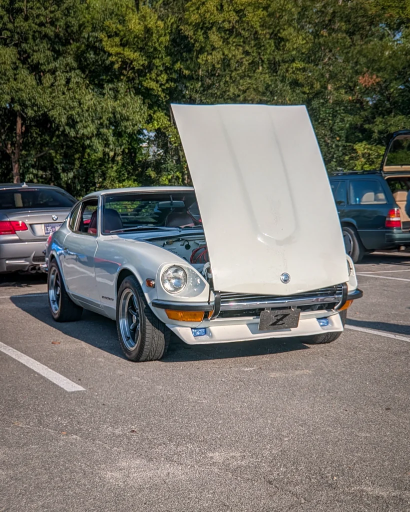 White Datsun 240Z with hood open in a parking lot at a car event during daytime