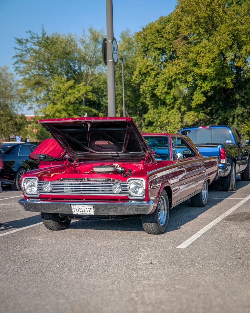 A red Plymouth Satellite with its hood raised sits in a parking lot among other vehicles at a car show.
