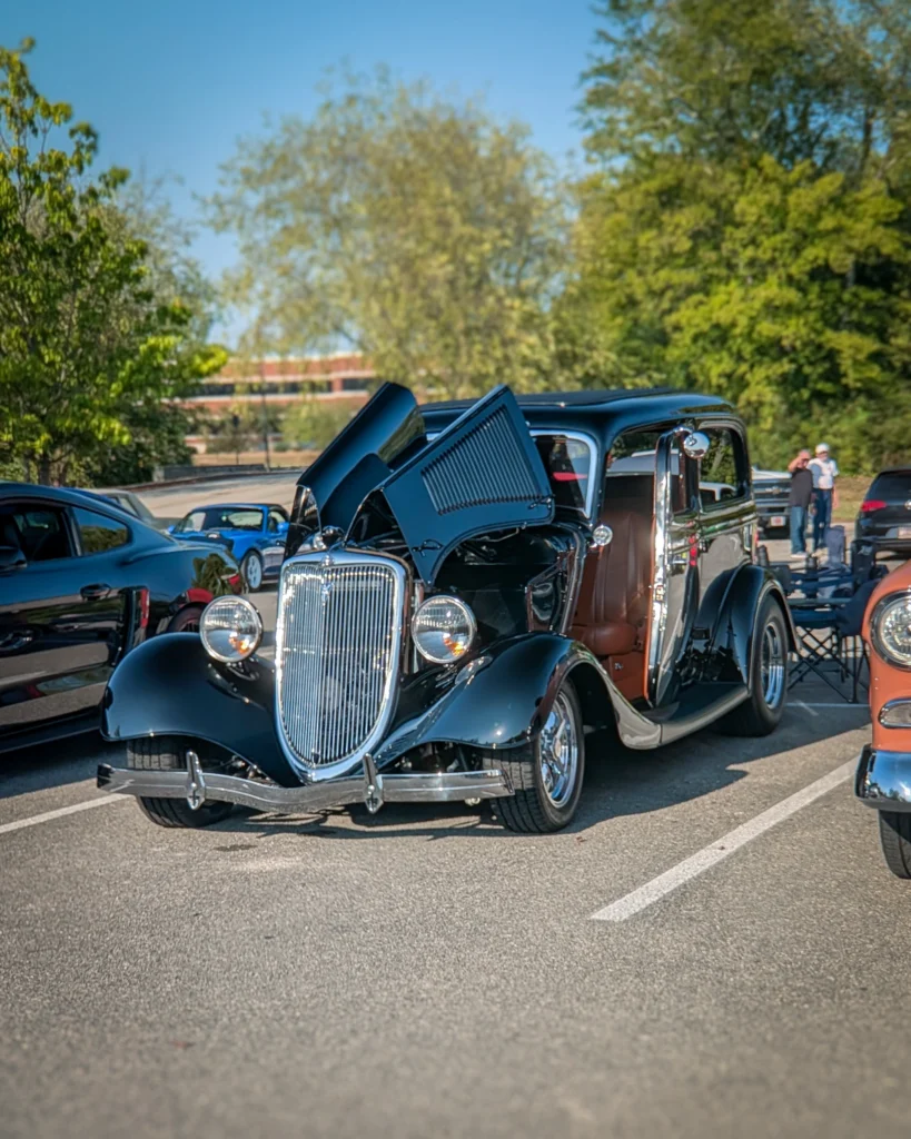 Black vintage Ford coupe with open hood parked in a lot surrounded by other vehicles and trees in the background