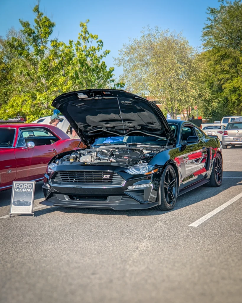 Black Ford Mustang with hood open showing engine at outdoor car event with other vehicles in background
