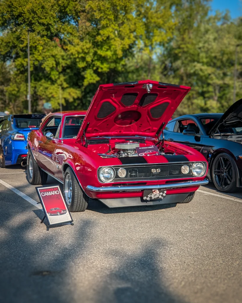 Red Chevrolet Camaro SS with hood open showing engine at outdoor car meet with other vehicles in background