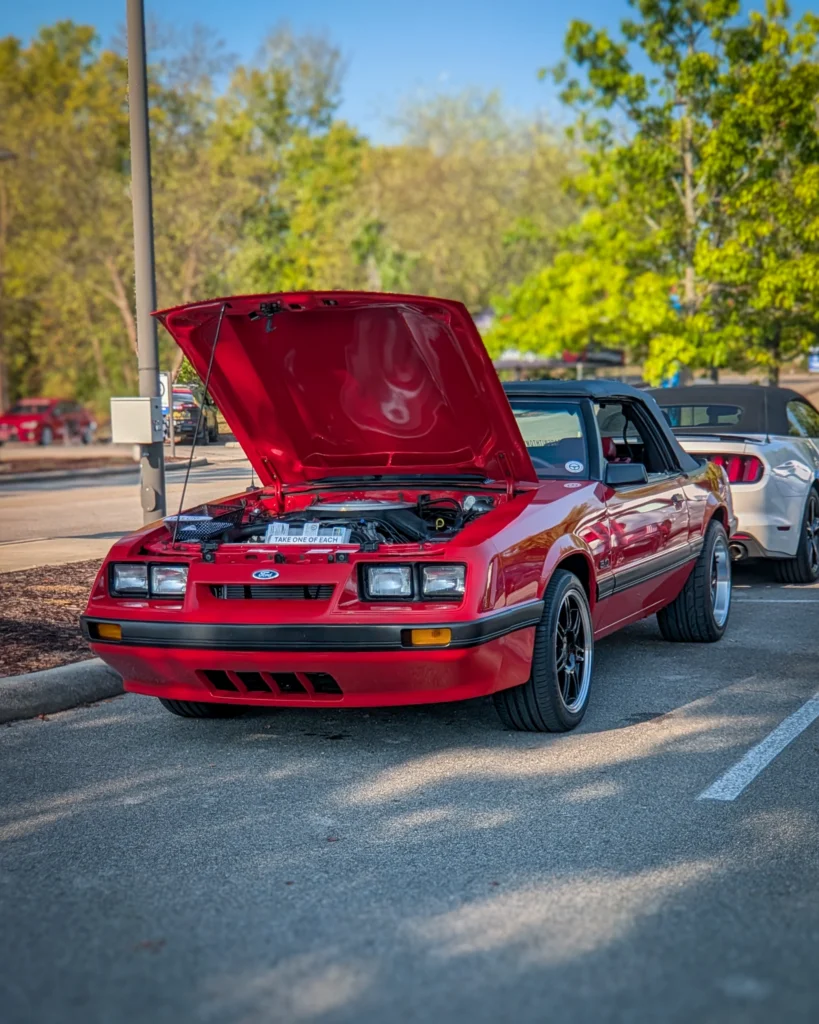 Red Ford Mustang convertible with hood open at an outdoor car event during daytime