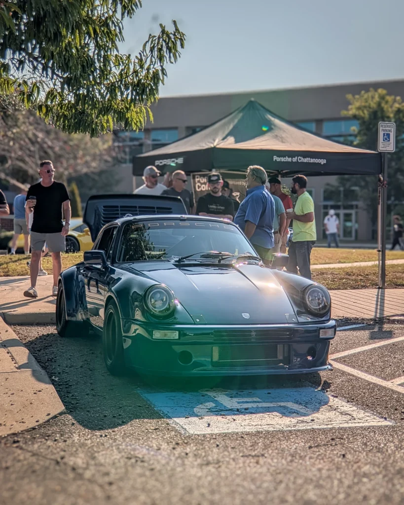 Dark green Porsche 911 parked in front of a Porsche of Chattanooga tent with several people standing nearby outdoors.
