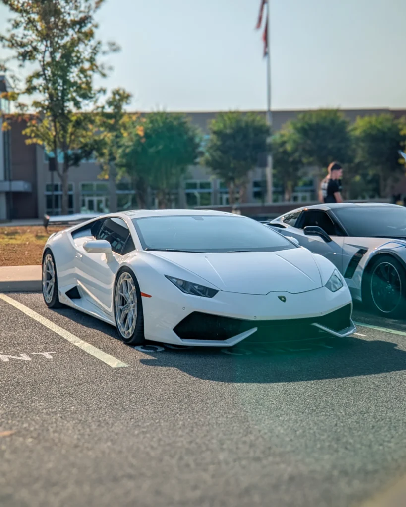 White Lamborghini Huracán parked next to a two-tone Chevrolet Corvette in an outdoor lot with trees and a building in the