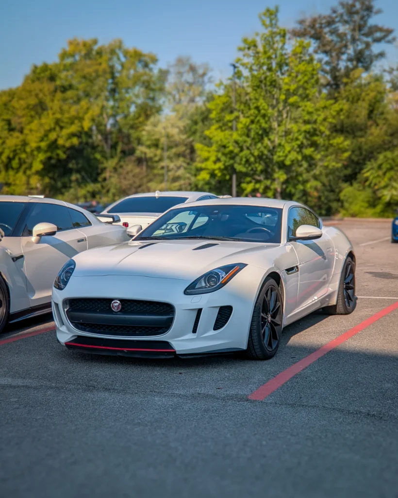 White Jaguar F-Type coupe parked diagonally in outdoor car event with other white vehicles and green trees in background