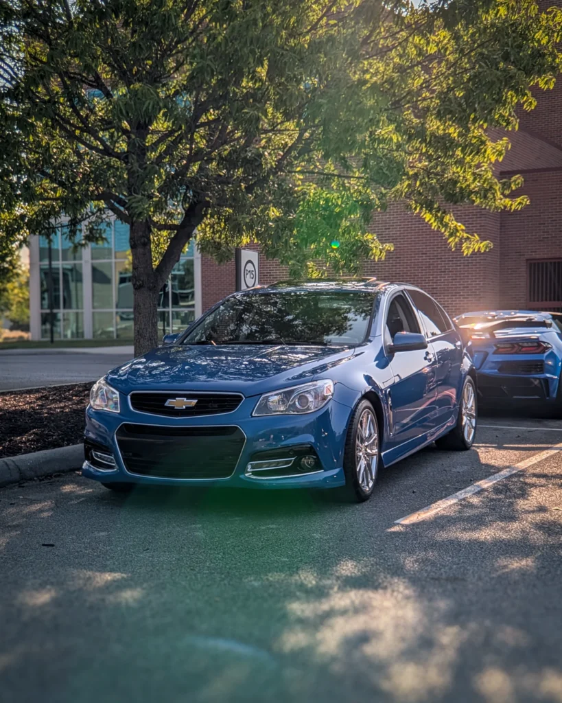 A blue Chevrolet SS is parked beneath a tree beside a brick building, viewed from the front driver's side.