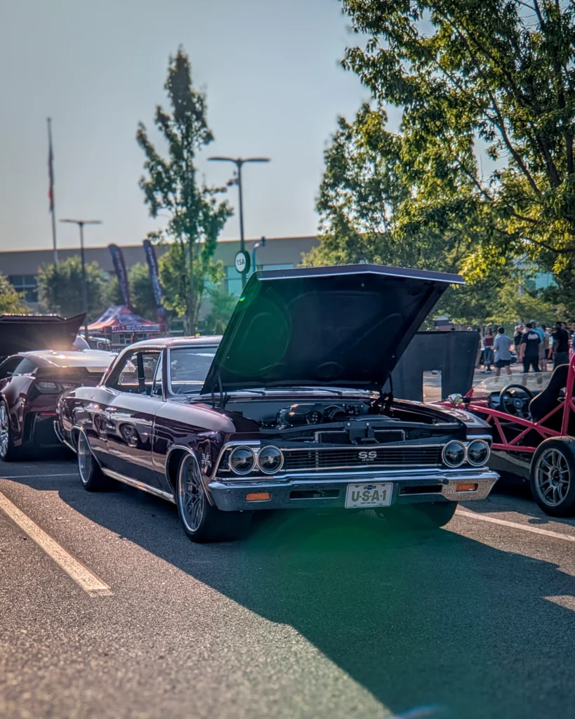 Maroon Chevrolet Chevelle SS 396 with hood open at an outdoor car event parking lot with other cars and people.