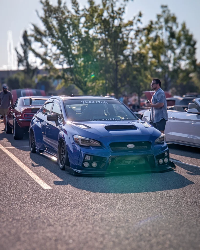 Blue Subaru WRX parked at an outdoor car event with other vehicles and people in the background under trees