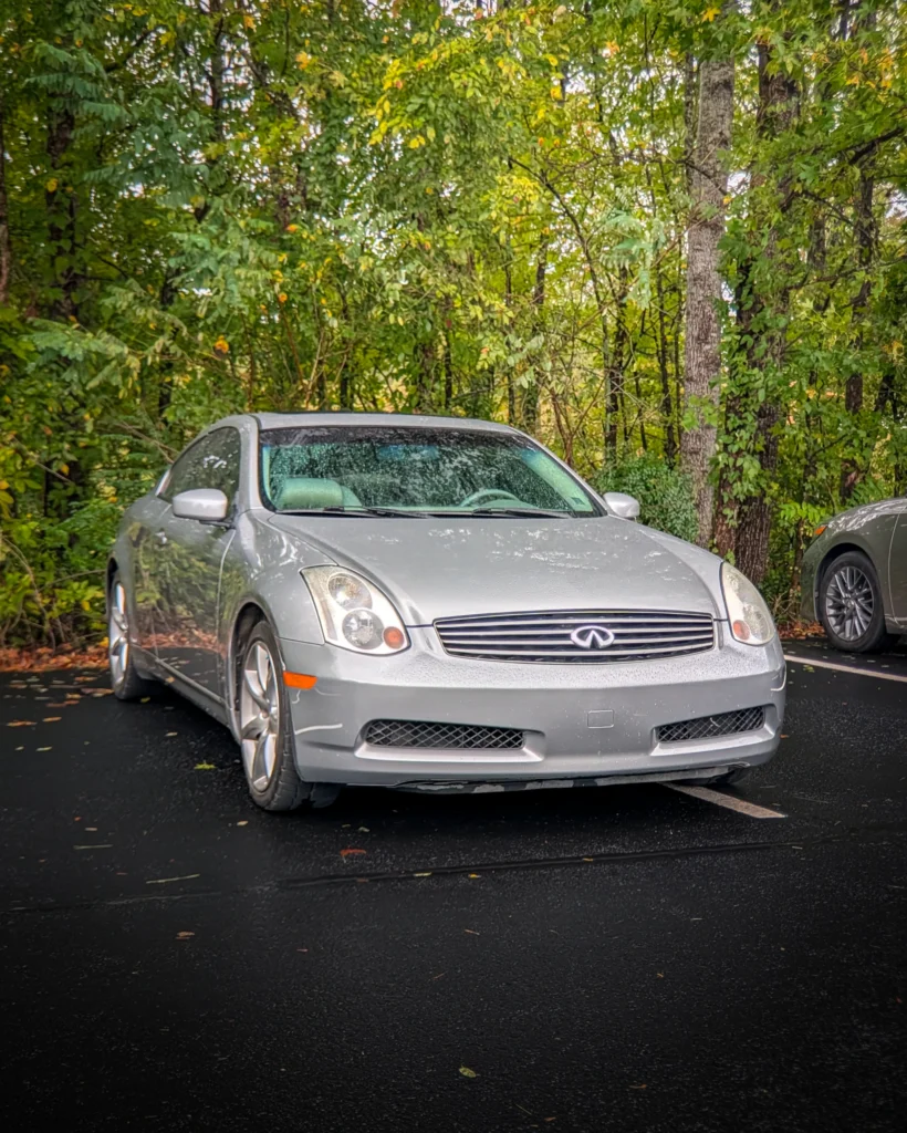 Silver Infiniti G35 coupe parked on wet asphalt with trees in background