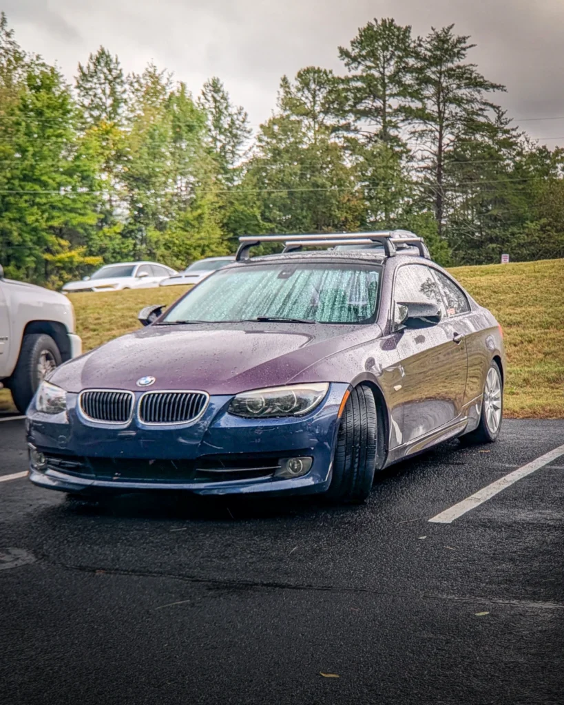 A blue BMW 3 Series coupe with roof racks is parked on wet pavement beside a grassy bank and trees.