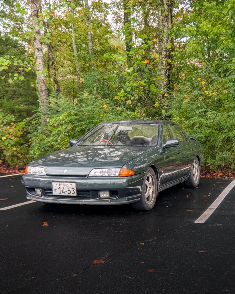 Dark green Nissan Skyline parked on wet asphalt near dense green foliage