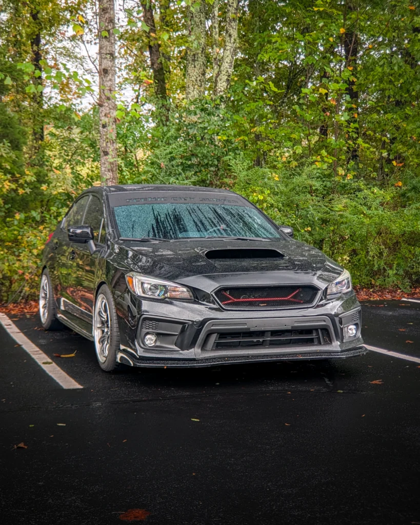Black Subaru WRX parked near a wooded area with moisture on the windshield and hood scoop visible