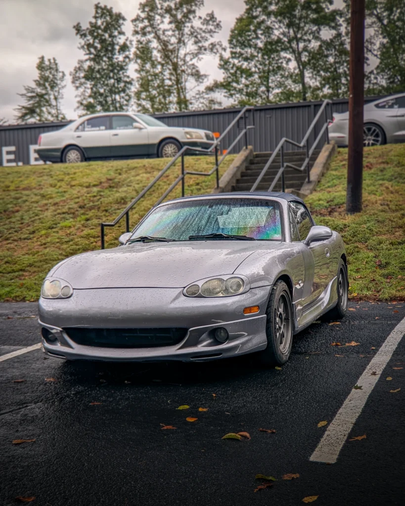 Silver Mazda MX-5 Miata convertible parked on wet pavement near a grassy slope with other parked cars in the background