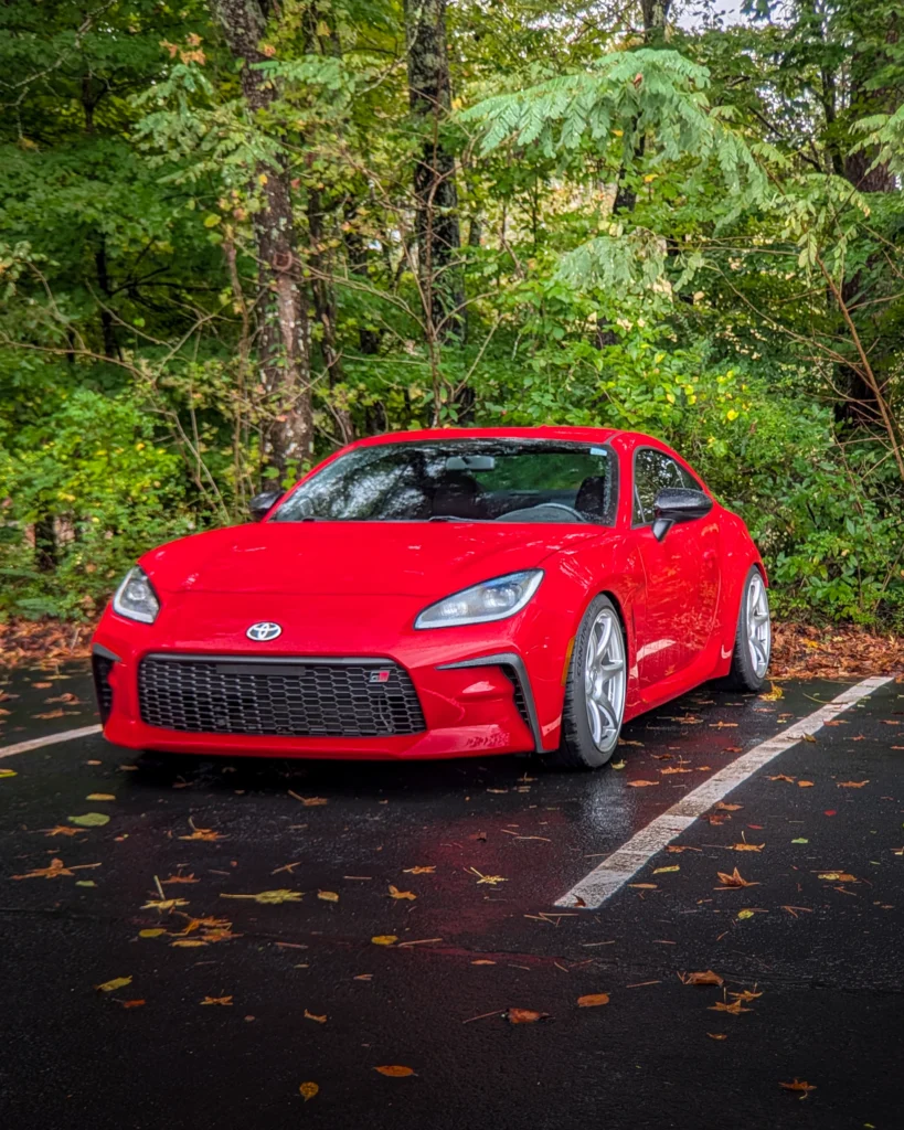 Red Toyota GR86 parked in forested lot on wet pavement with fallen leaves scattered