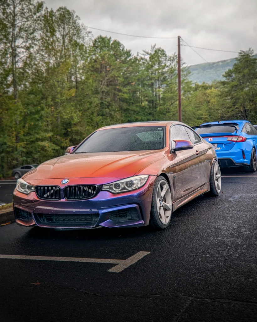 Orange and purple BMW 4-Series parked on asphalt with a blue Honda hatchback behind it and trees in the background